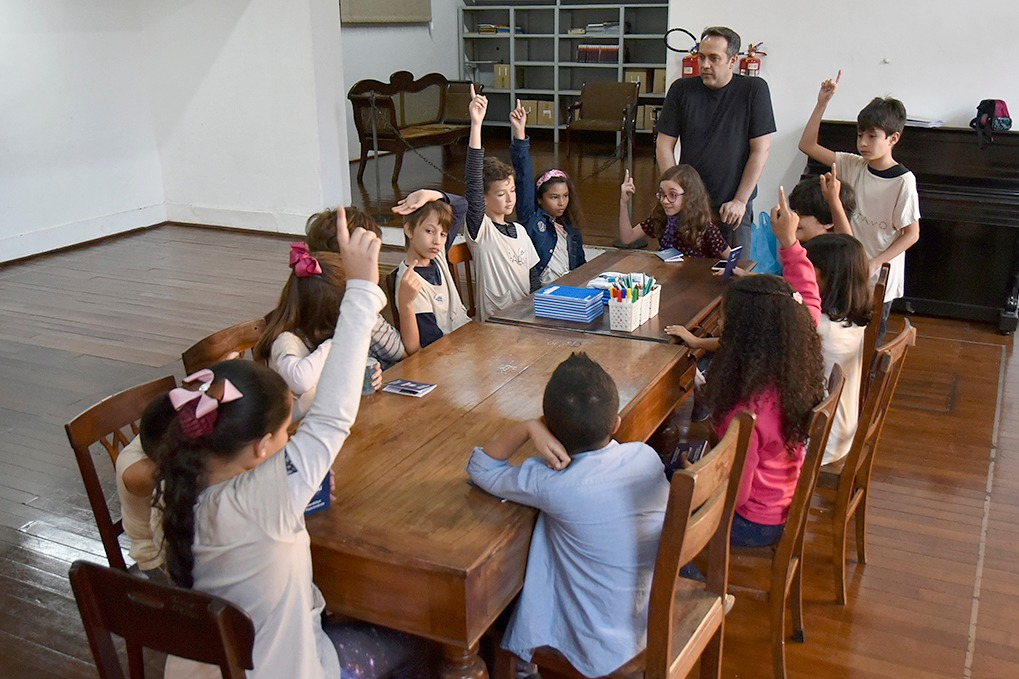 Mesa de madeira, com crianças sentadas ao redor, algumas com as mãos levantadas, enquanto atendem a instruções de homem em pé na cabeceira da mesa