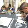 Crianças pintam com pincel placas de madeira, sobre mesa no FAB LAB