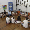 Crianças sentadas em círculo no chão de madeira, com relógios diversos na parede