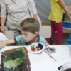 Crianças fazendo atividades de pintura com aquarela, com responsáveis e oficineira ao fundo