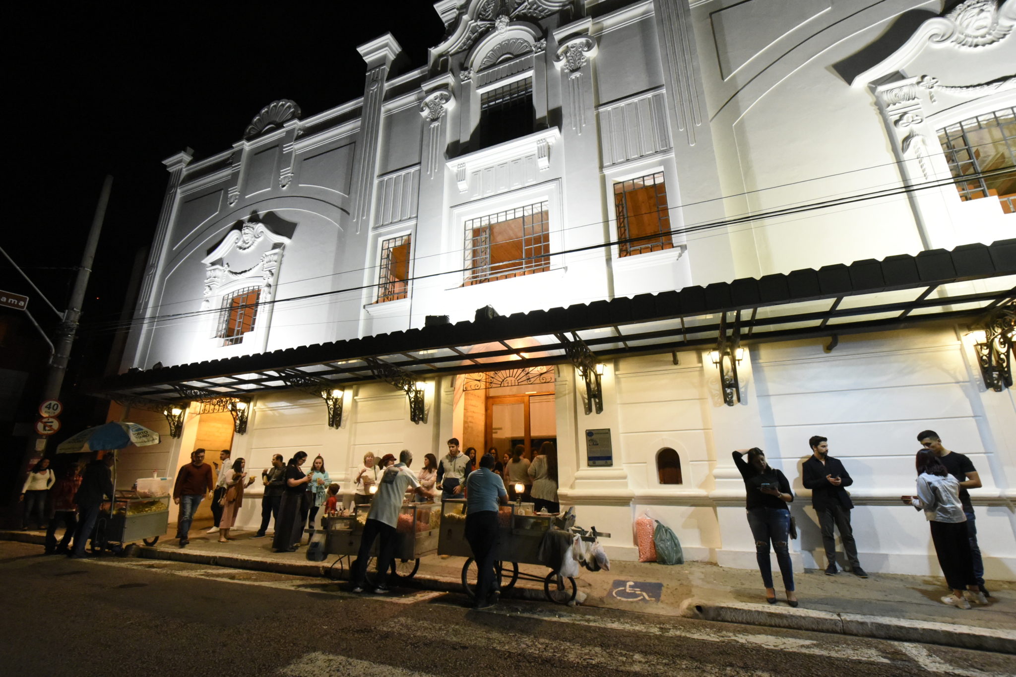 Foto noturna da fachada do teatro Polytheama, com pessoas aguardando para entrar