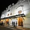 Foto noturna do teatro, com fachada iluminada e pessoas na calçada