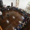 Foto vista do alto, em sala com chão de madeira, e grupo sentado em semicírculo, voltado para homem que dá a palestra