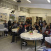 Sala de aula, com mesas espalhadas, e pessoas sentadas em cadeiras, assistindo a uma apresentação