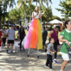 Adultos e crianças acompanham intervenção artística, com mulher andando de perna de pau