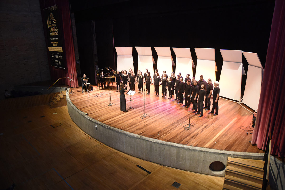 Palco de teatro, com cantores com roupas pretas perfilados, com regente de costas e pessoa ao piano ao fundo